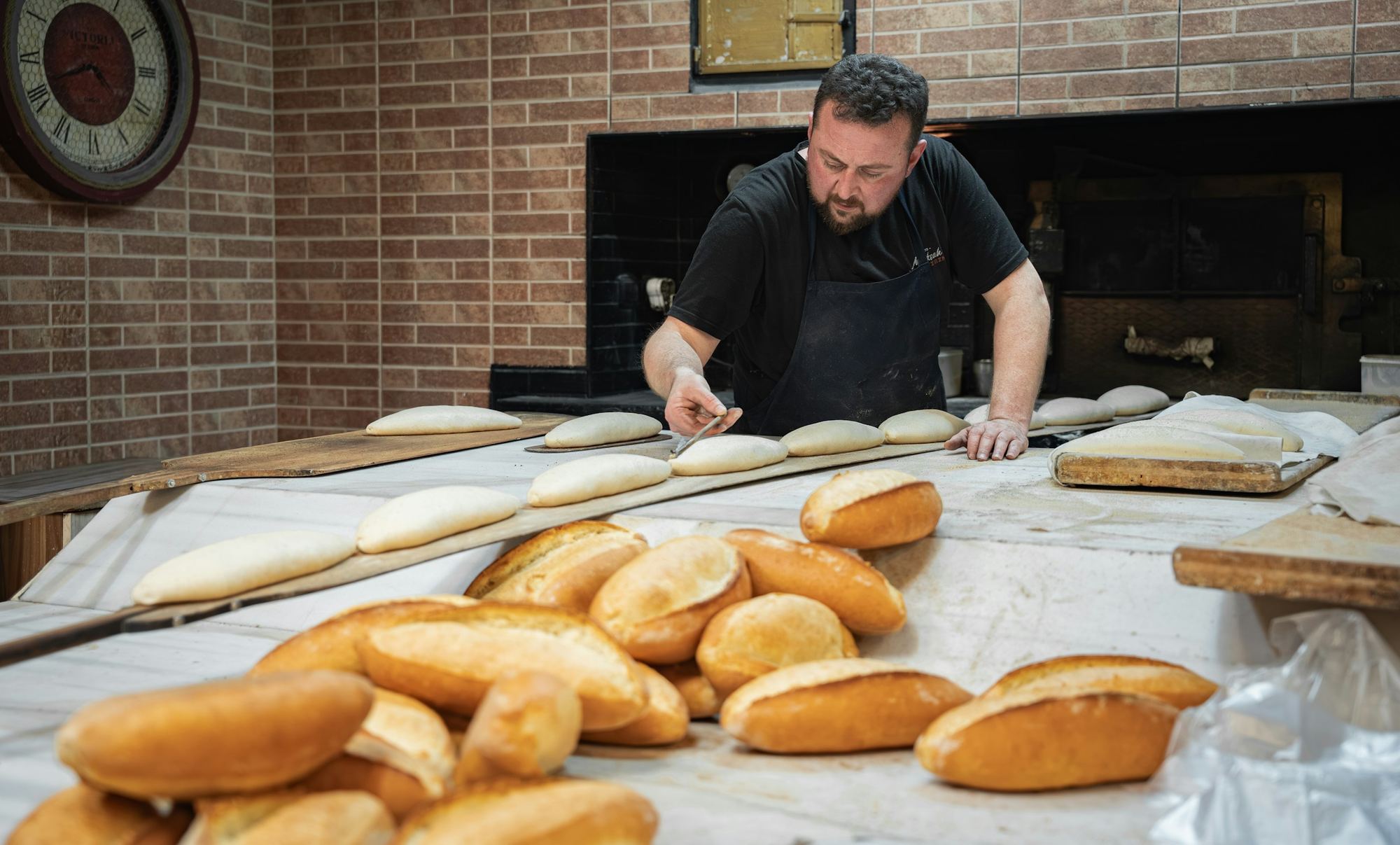 Panadero trabajando en su panadería con horno de ladrillo, con panes en primer plano.