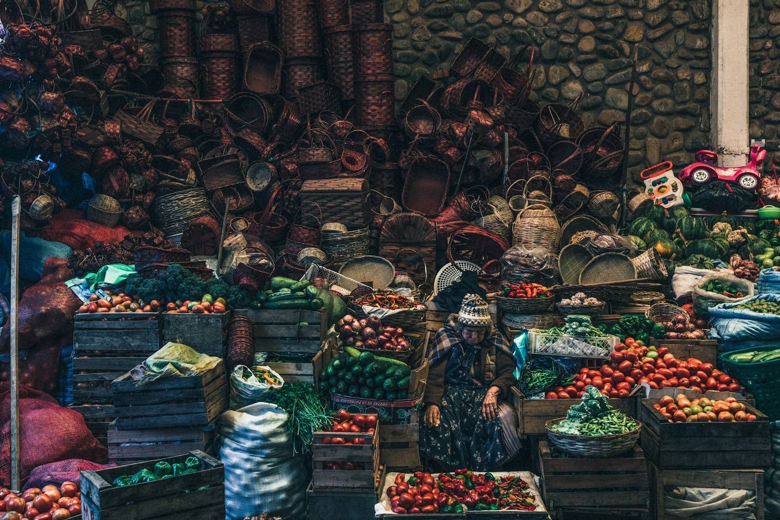 Puesto de mercado andino con cajones de verduras, canastos tejidos y una vendedora.